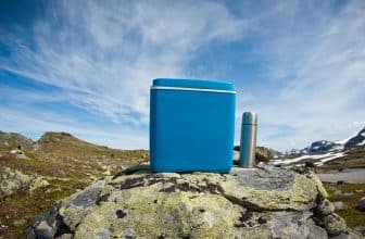 Graue Thermoskanne neben einer blauen Kühlbox auf einem Fels in freier Landschaft.